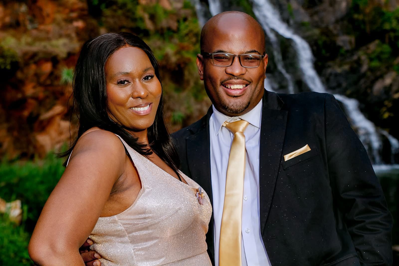 The couple smiling side by side in formal wear, a waterfall behind them