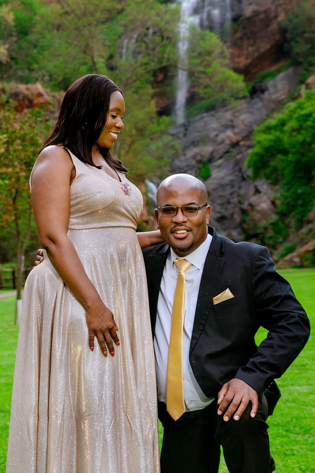 He kneels on one knee in a dark suit and gold tie as she, in a champagne gown, smiles down at him, a waterfall behind