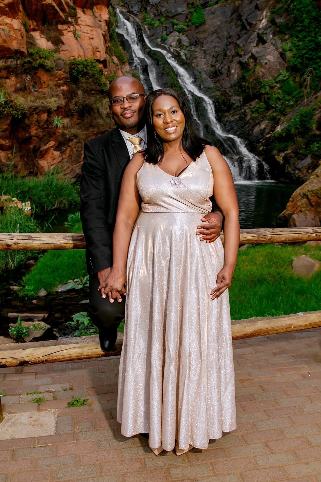 The couple standing close in front of a waterfall, he in a dark suit and gold tie, she in a floor-length champagne gown